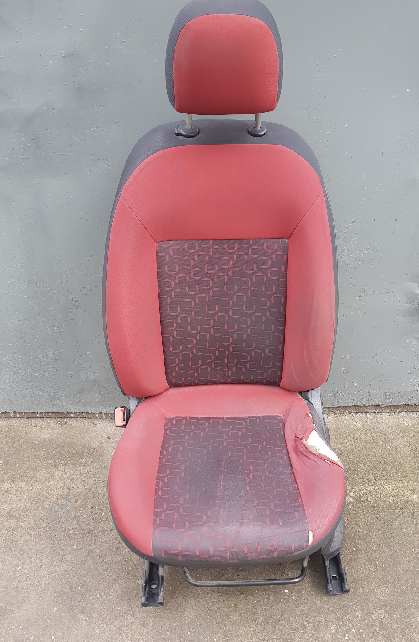 Czerwono-szary fotel samochodowy z widocznym przetarciem na siedzisku, na tle szarej ściany i betonowej podłogi.