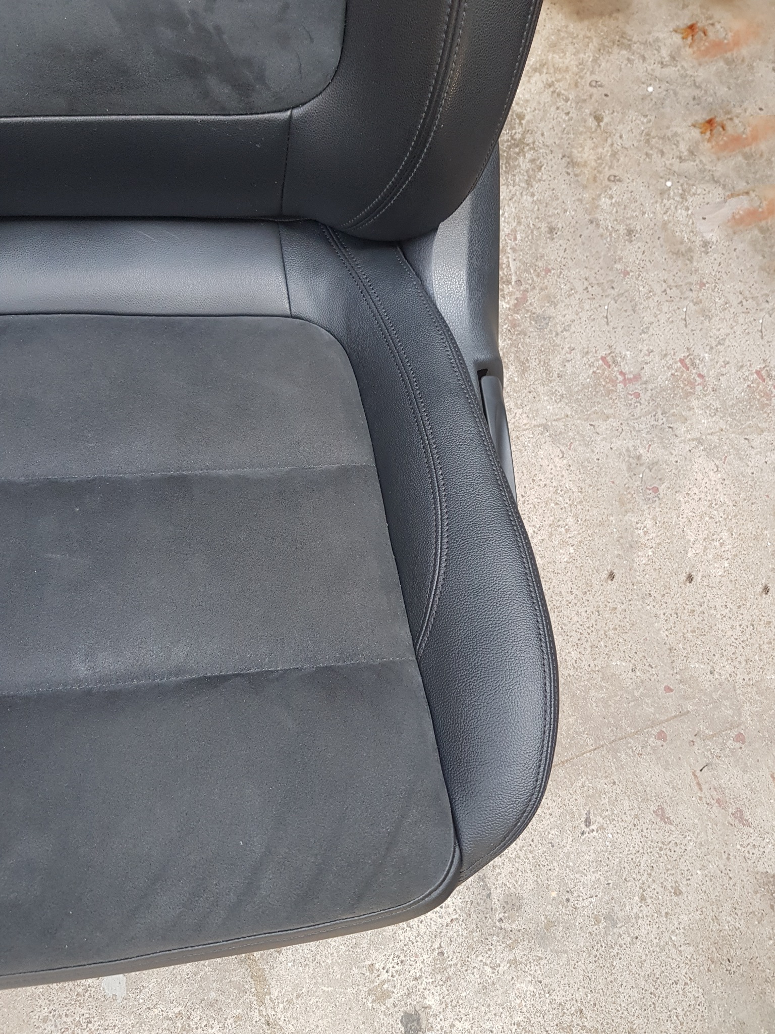 Fragment czarnego fotela samochodowego z elementami z alcantary, widoczne szwy i faktura materiałów, na tle betonowej posadzki.