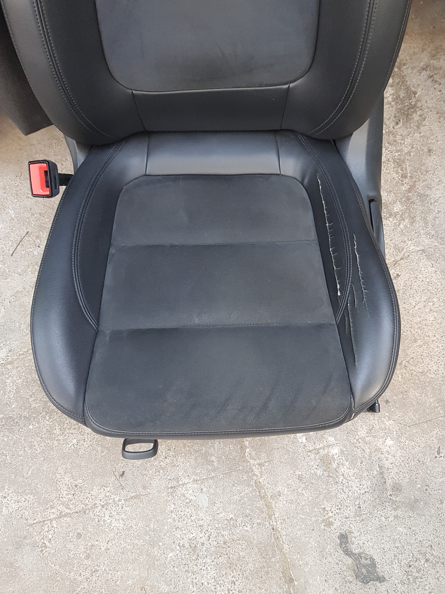 Czarne, zniszczone skórzane siedzenie samochodowe z widocznymi pęknięciami i zużyciem na siedzisku, sfotografowane na szarym betonowym podłożu.