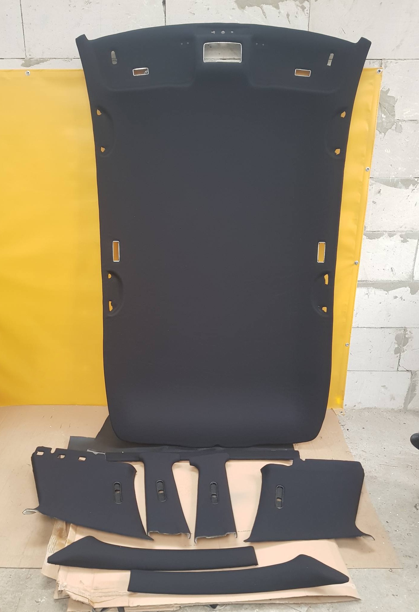 Zdemontowany, czarny podsufitka samochodowa i elementy tapicerki słupków na brązowym kartonie, z żółtym tłem, widoczne mocowania i otwory.