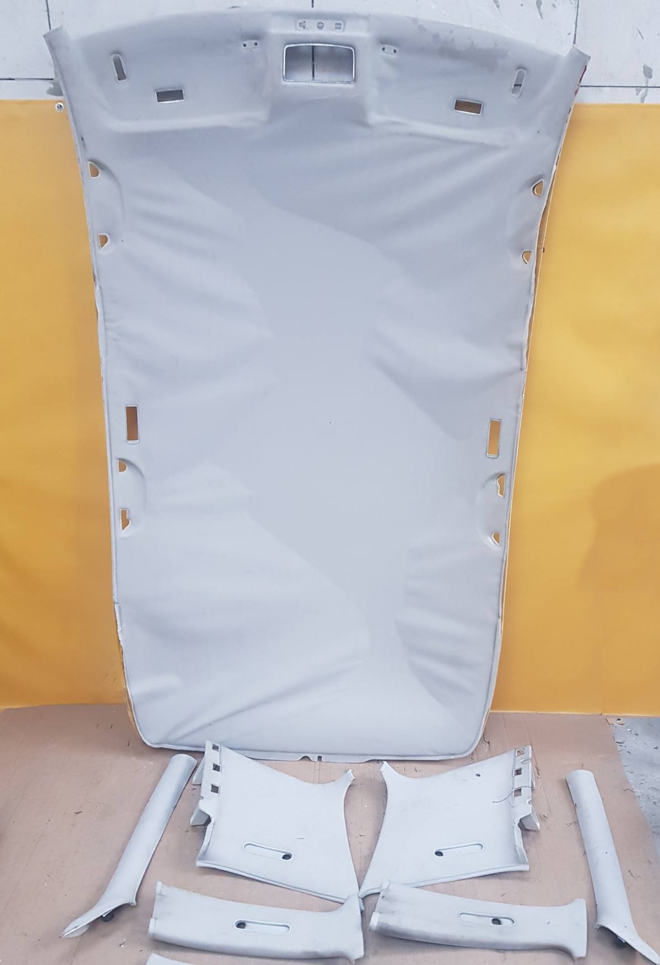 Wnętrze samochodu zdemontowane do tapicerowania: podsufitka i elementy słupków w jasnym odcieniu szarości, widoczne otwory montażowe i detale wykończeniowe.