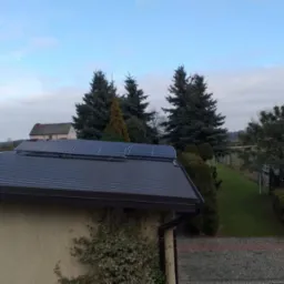 Panele słoneczne na metalowym dachu domu jednorodzinnego, widok z góry na tle zieleni i pochmurnego nieba.