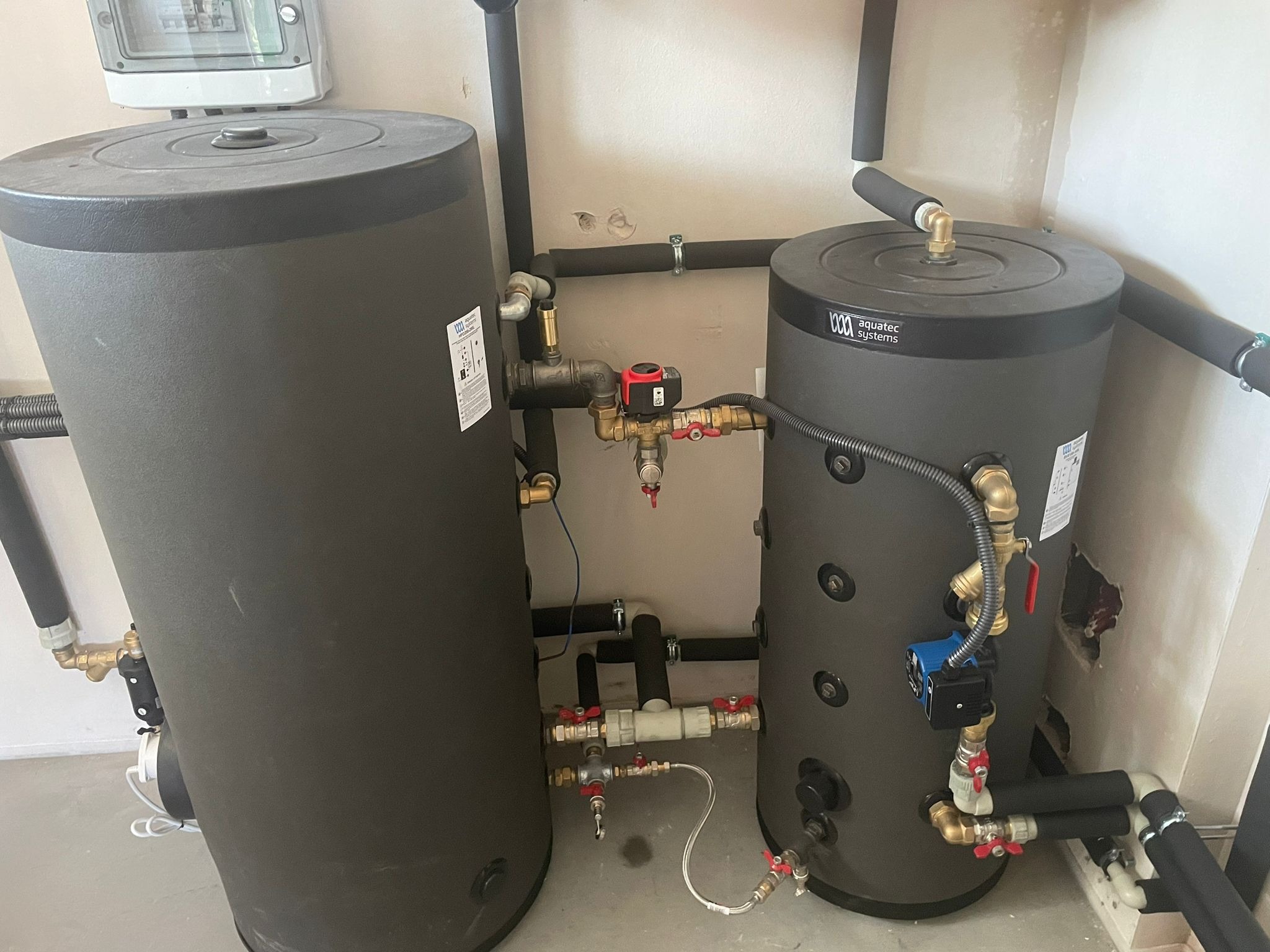 Dwa szare zbiorniki akumulacyjne ciepłej wody użytkowej z licznymi rurami i zaworami, instalacja w pomieszczeniu technicznym z widocznymi śladami po wierceniu w ścianie.
