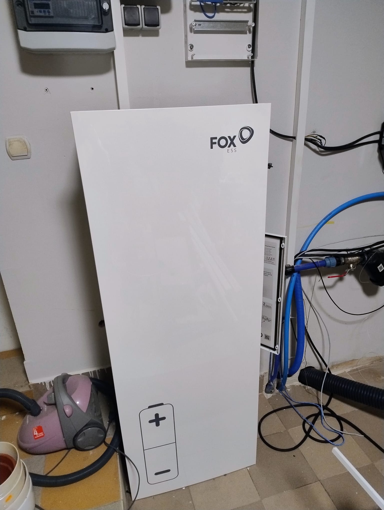 Biały moduł bateryjny Fox ESS z ikoną baterii i logiem firmy, widoczny na tle instalacji elektrycznej i hydraulicznej w pomieszczeniu gospodarczym, obok odkurzacza i zabrudzonej podłogi.