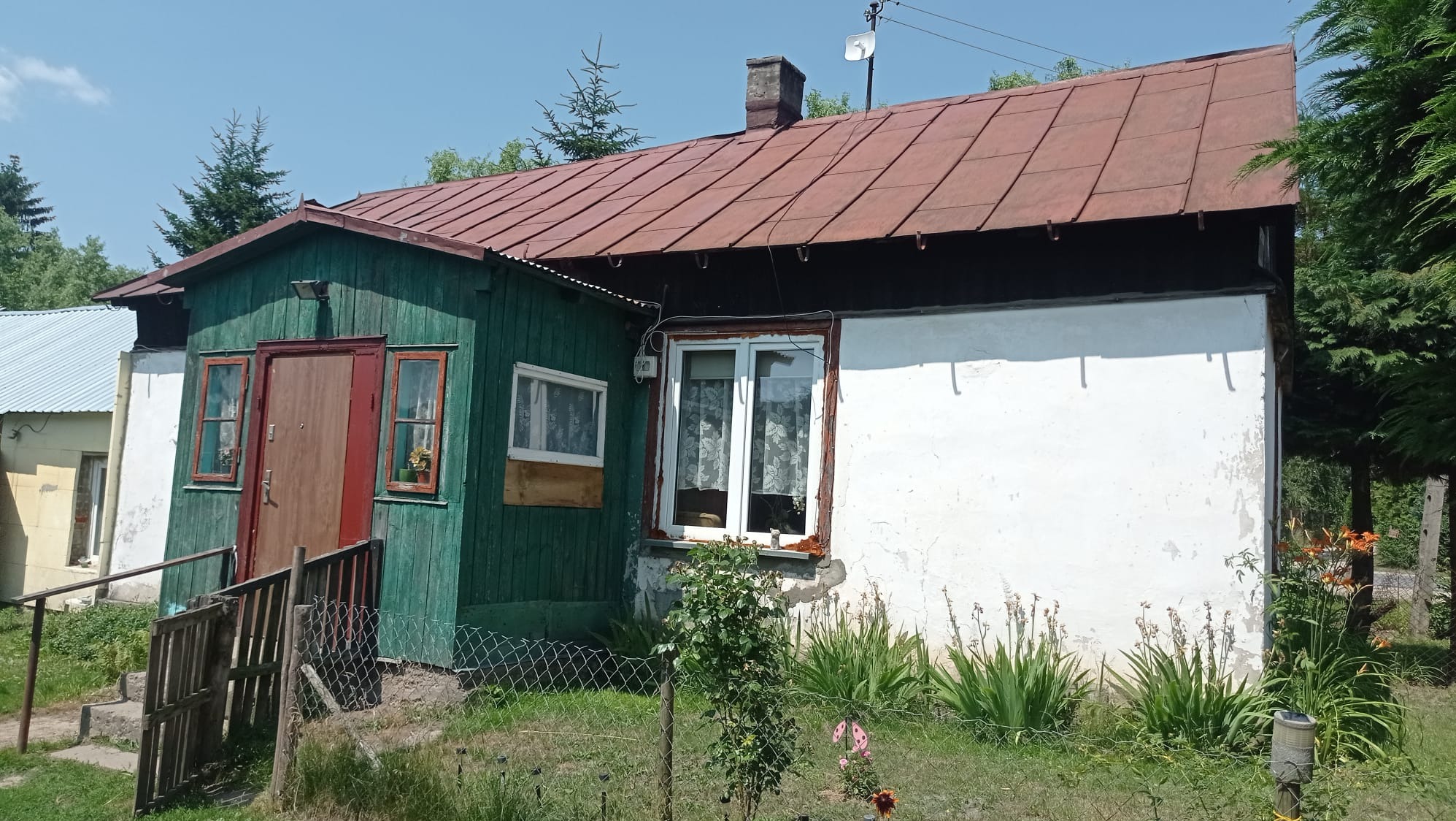 Starszy, częściowo ocieplony dom z zieloną drewnianą werandą, białą ścianą i rdzawym dachem, otoczony zielenią i ogrodzeniem z siatki.