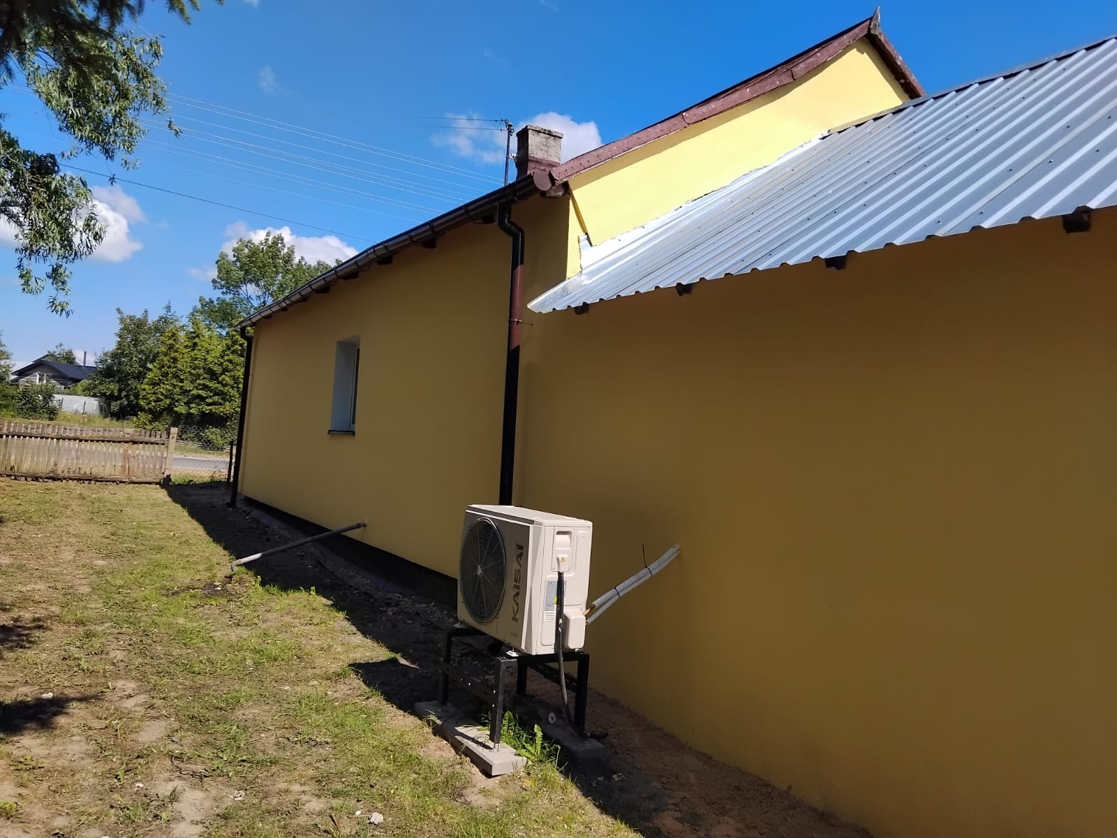 Żółty dom jednorodzinny z nową, metalową blachodachówką i zamontowanym klimatyzatorem Kaisai na zewnątrz, widok z boku w słoneczny dzień.