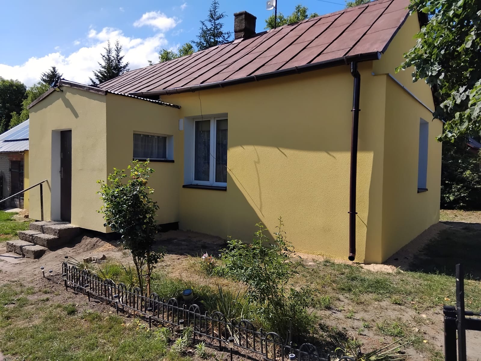 Żółty dom jednorodzinny po ociepleniu elewacji, widoczne brązowe rynny i białe okna, ogródek z niskim metalowym płotkiem.