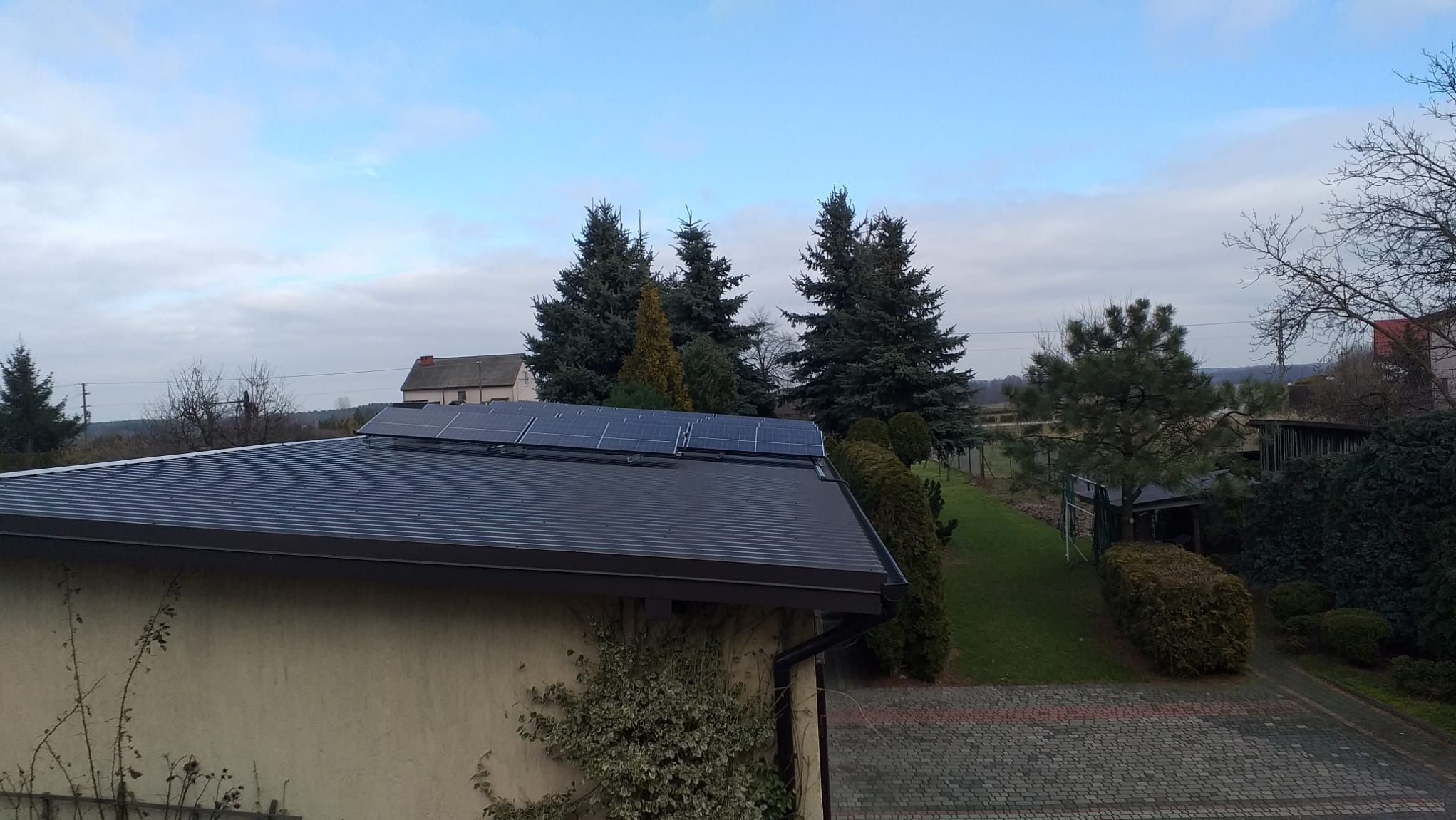 Panele słoneczne na metalowym dachu domu jednorodzinnego, widok z góry na tle zieleni i pochmurnego nieba.