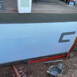 Przedsiębiorstwo Usługowo Handlowe JACEK CHMIELEWSKI - Ocieplenie fundamentu budynku płytami styropianowymi z widocznym fragmentem rury odprowadzającej wodę deszczową i narzędziami budowlanymi w ziemi