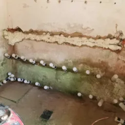 Wilgotna ściana w trakcie osuszania z widocznymi nawiertami i pianą izolacyjną w szczelinie, oraz rzędem plastikowych rurek z etykietami w otworach, na podłodze resztki wody i sprzęt budowlany.