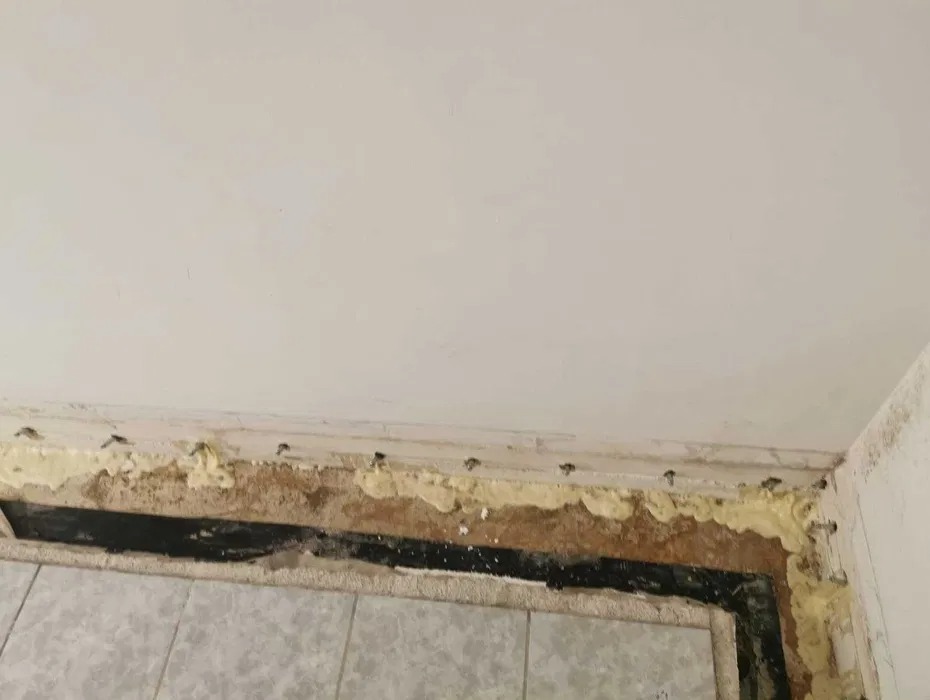 Fragment ściany z widoczną warstwą izolacyjną z pianki montażowej i czarnej folii, zabezpieczoną kołkami, nad płytkami podłogowymi.