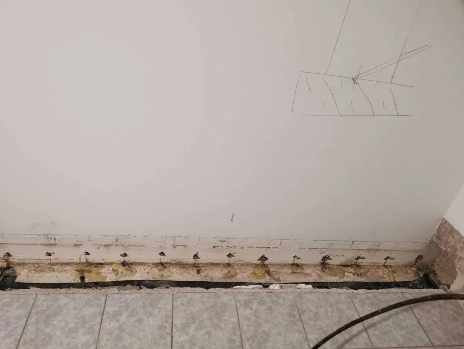 Przygotowany fragment ściany przy podłodze, z widocznymi otworami i elementami uszczelniającymi, prawdopodobnie przed nałożeniem warstwy izolacyjnej. Na ścianie widoczne prowizoryczne oznaczenia...