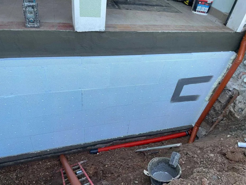 Ocieplenie fundamentu budynku płytami styropianowymi z widocznym fragmentem rury odprowadzającej wodę deszczową i narzędziami budowlanymi w ziemi