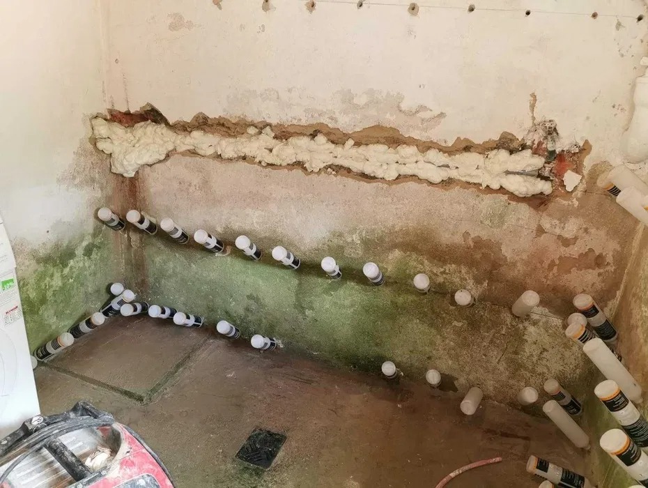 Wilgotna ściana w trakcie osuszania z widocznymi nawiertami i pianą izolacyjną w szczelinie, oraz rzędem plastikowych rurek z etykietami w otworach, na podłodze resztki wody i sprzęt budowlany.