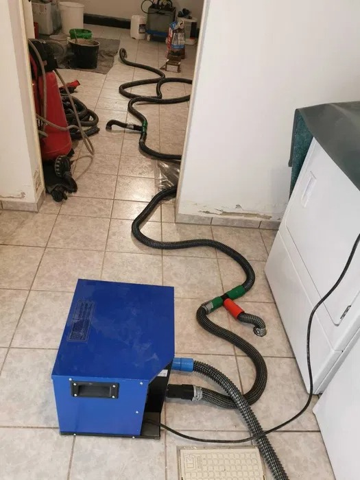 Profesjonalny sprzęt do usuwania wilgoci z pomieszczenia, z długimi wężami rozłożonymi na kafelkowej podłodze, widoczna pralka i ściana z uszkodzeniami.