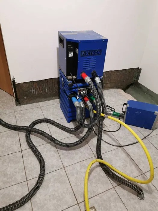 Niebieskie urządzenie FIXTRON z podłączonymi przewodami w pomieszczeniu z uszkodzoną ścianą przy podłodze, prawdopodobnie podczas procesu usuwania wilgoci.