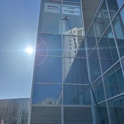 MCSTEAM SPÓŁKA Z OGRANICZONĄ ODPOWIEDZIALNOŚCIĄ - Mycie okien na wysokości w nowoczesnym biurowcu z użyciem specjalistycznego sprzętu. Widoczne smugi wody i odbicie otoczenia w czystych szybach.