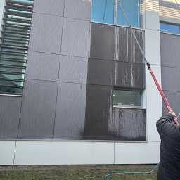 MCSTEAM SPÓŁKA Z OGRANICZONĄ ODPOWIEDZIALNOŚCIĄ - Mycie okien na wysokości w budynku biurowym. Widoczny fragment elewacji z umytymi i brudnymi oknami oraz pracownik z lancą teleskopową. W tle trawnik i niebieski wąż.