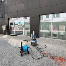 MCSTEAM SPÓŁKA Z OGRANICZONĄ ODPOWIEDZIALNOŚCIĄ - Profesjonalny sprzęt do mycia elewacji i okien na zewnątrz budynku z szarą kostką brukową. Niebieski wąż na wózku i pomarańczowe kable.