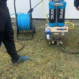 MCSTEAM SPÓŁKA Z OGRANICZONĄ ODPOWIEDZIALNOŚCIĄ - Profesjonalny sprzęt do mycia okien Ionic Systems z bębnem na wąż, na trawie. Widoczne filtry, manometr i złącza. Częściowo widoczne nogi osoby obsługującej.