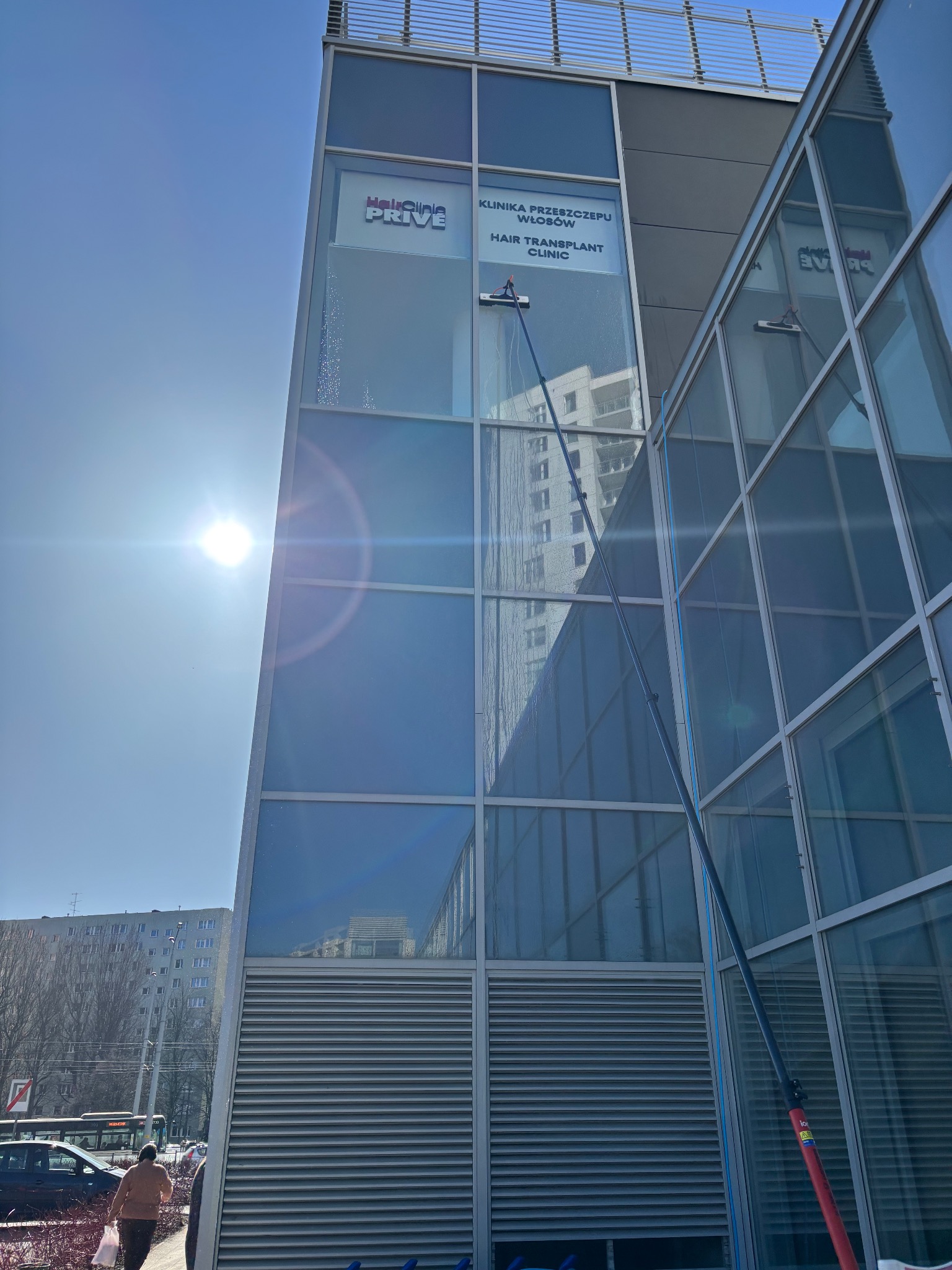 Mycie okien na wysokości w nowoczesnym biurowcu z użyciem specjalistycznego sprzętu. Widoczne smugi wody i odbicie otoczenia w czystych szybach.