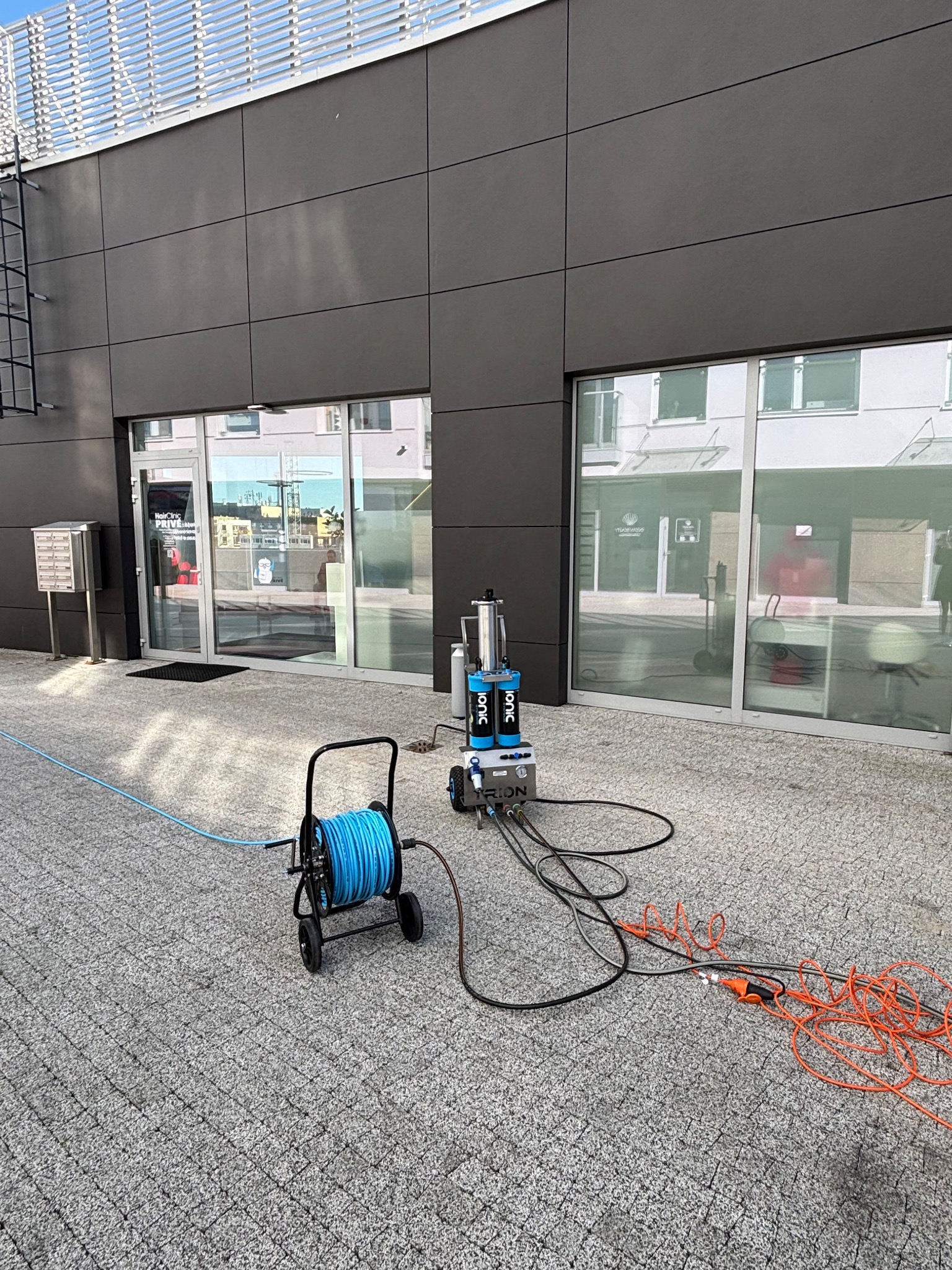 Profesjonalny sprzęt do mycia elewacji i okien na zewnątrz budynku z szarą kostką brukową. Niebieski wąż na wózku i pomarańczowe kable.