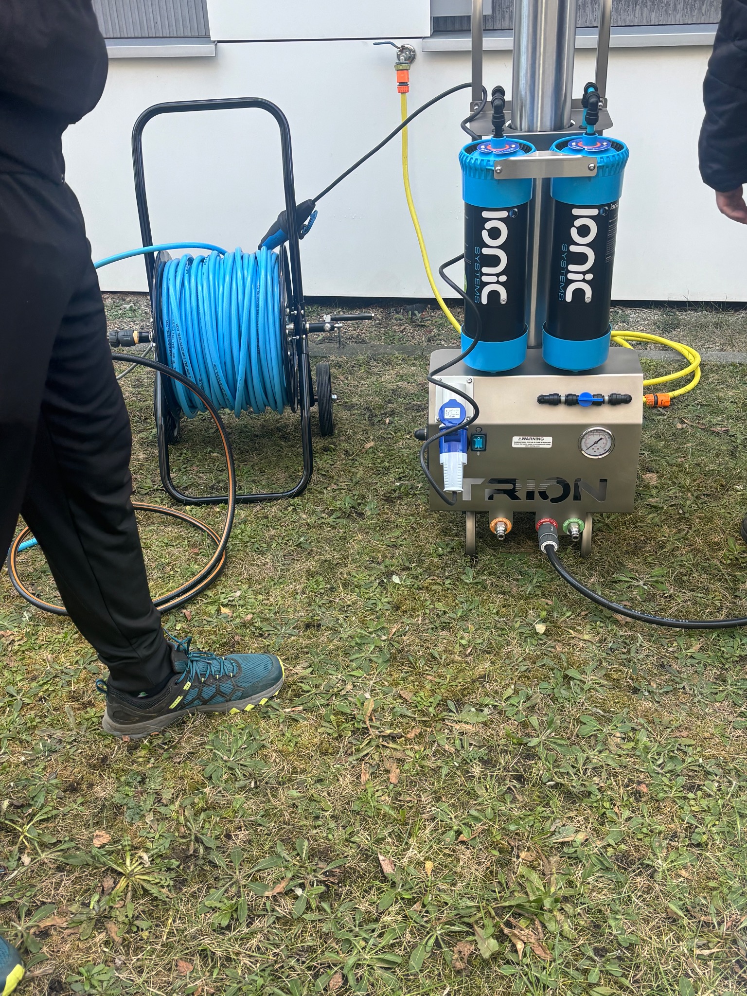 Profesjonalny sprzęt do mycia okien Ionic Systems z bębnem na wąż, na trawie. Widoczne filtry, manometr i złącza. Częściowo widoczne nogi osoby obsługującej.
