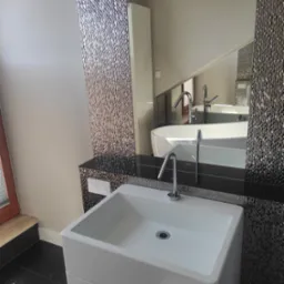 Nowoczesna łazienka z białą umywalką na tle mozaiki i lustra odbijającego wannę. Minimalistyczny design, czarne płytki podłogowe, lampa sufitowa.