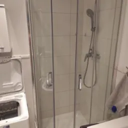 Wnętrze łazienki po sprzątaniu: kabina prysznicowa z chromowanymi detalami, pralka z otwartymi drzwiczkami i umywalka z ręcznikami. Czyste płytki na ścianach.