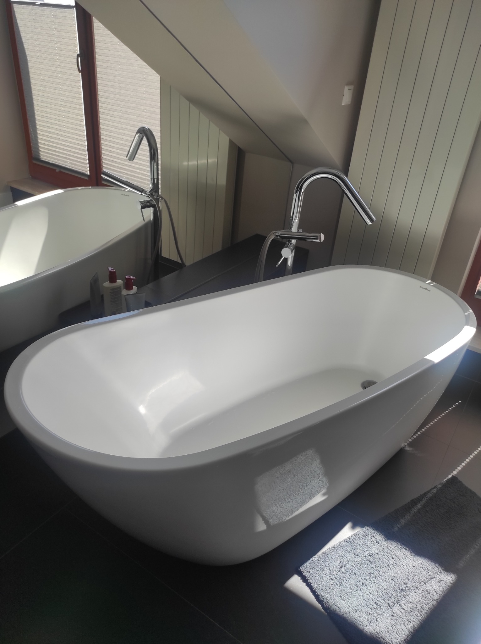 Biała, wolnostojąca wanna w nowoczesnej łazience na poddaszu z oknem. Widoczna armatura, kosmetyki i odbicie w lustrze. Czysta i zadbana.