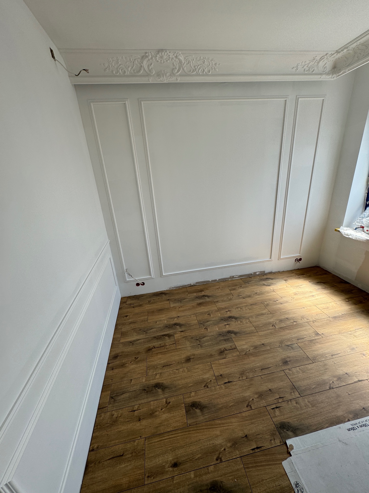 Pokój po remoncie z drewnopodobną podłogą, białe ściany z dekoracyjnymi listwami i rozetą sufitową, widoczne niedokończone gniazdka elektryczne i karton na podłodze.