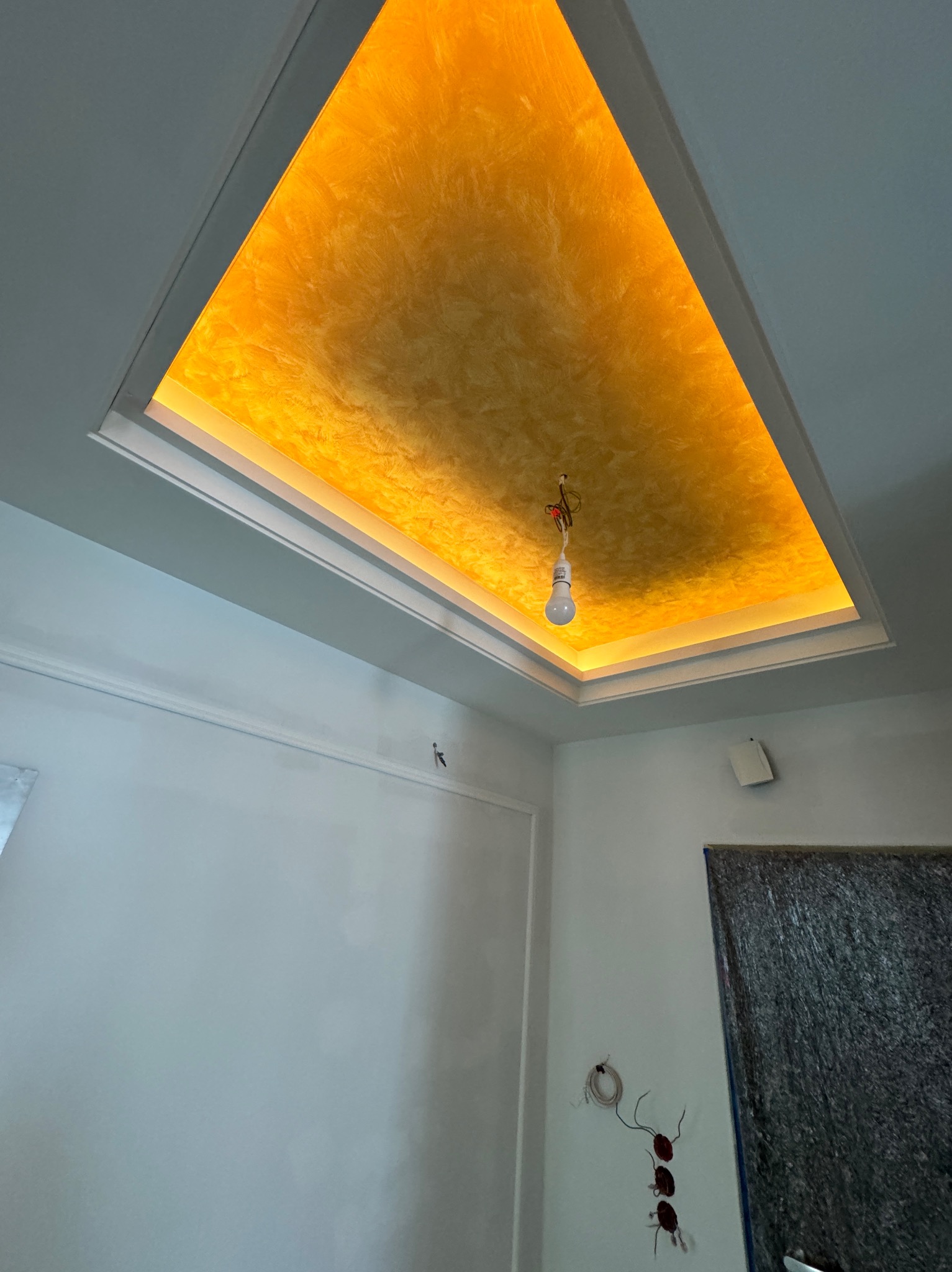 Podwieszany sufit z oświetleniem LED i dekoracyjną fakturą w odcieniach złota, widoczna wisząca żarówka i ślady po instalacji elektrycznej na ścianie.
