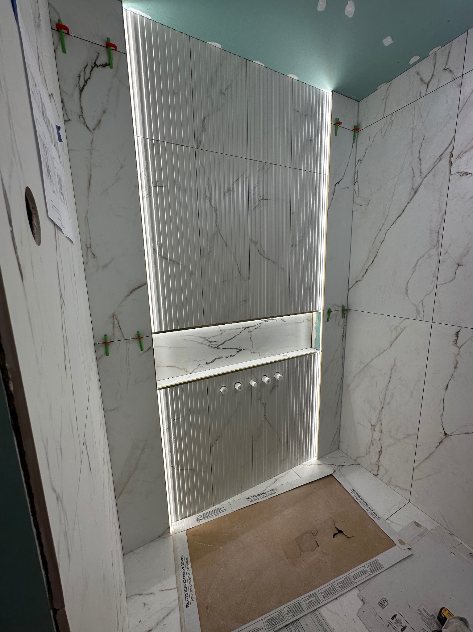 Wnętrze łazienki w trakcie kładzenia płytek imitujących marmur, z podświetlaną wnęką z półką i czterema wystającymi elementami armatury, widoczne klipsy poziomujące płytki.