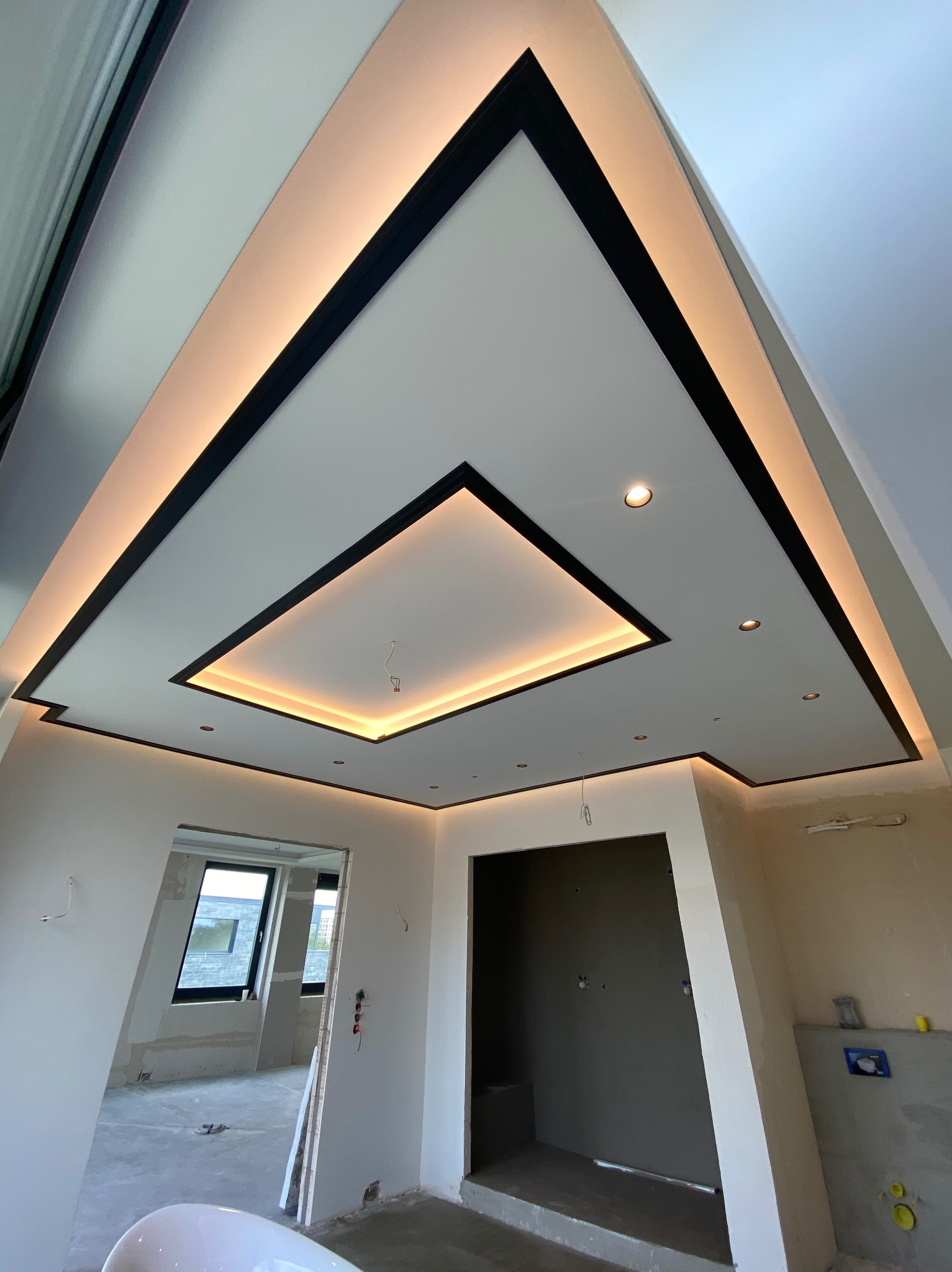 Sufit podwieszany z czarnymi obramowaniami i oświetleniem LED w pomieszczeniu w trakcie prac wykończeniowych; widoczne niedokończone ściany i instalacje.