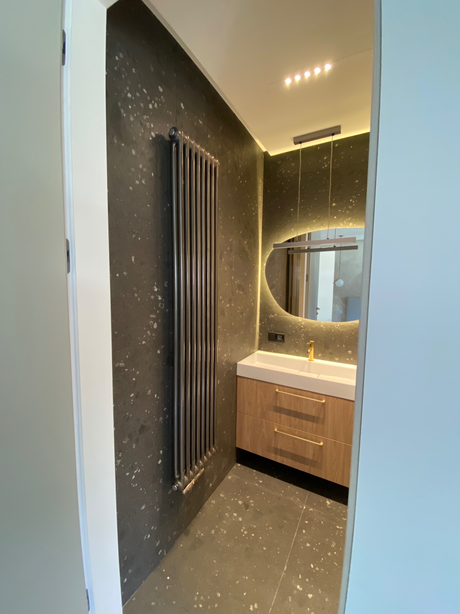 Nowoczesna łazienka z czarną ścianą w plamki, grzejnikiem drabinkowym, szafką pod umywalkę z drewnopodobnymi frontami i podświetlanym lustrem owalnym.