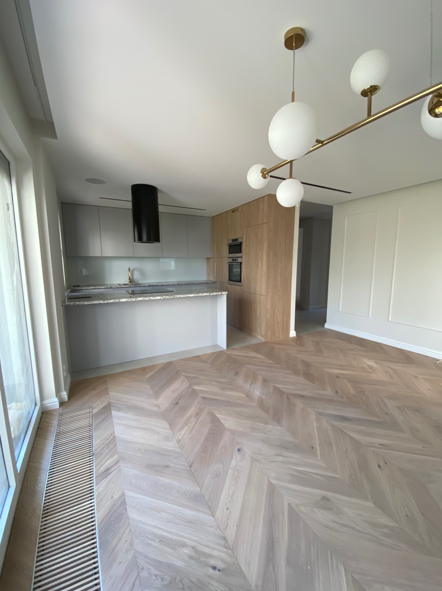 Nowoczesna kuchnia z drewnianą podłogą ułożoną w jodełkę, szare szafki i czarny okap, oświetlona designerską lampą z białymi kulami i złotymi elementami.