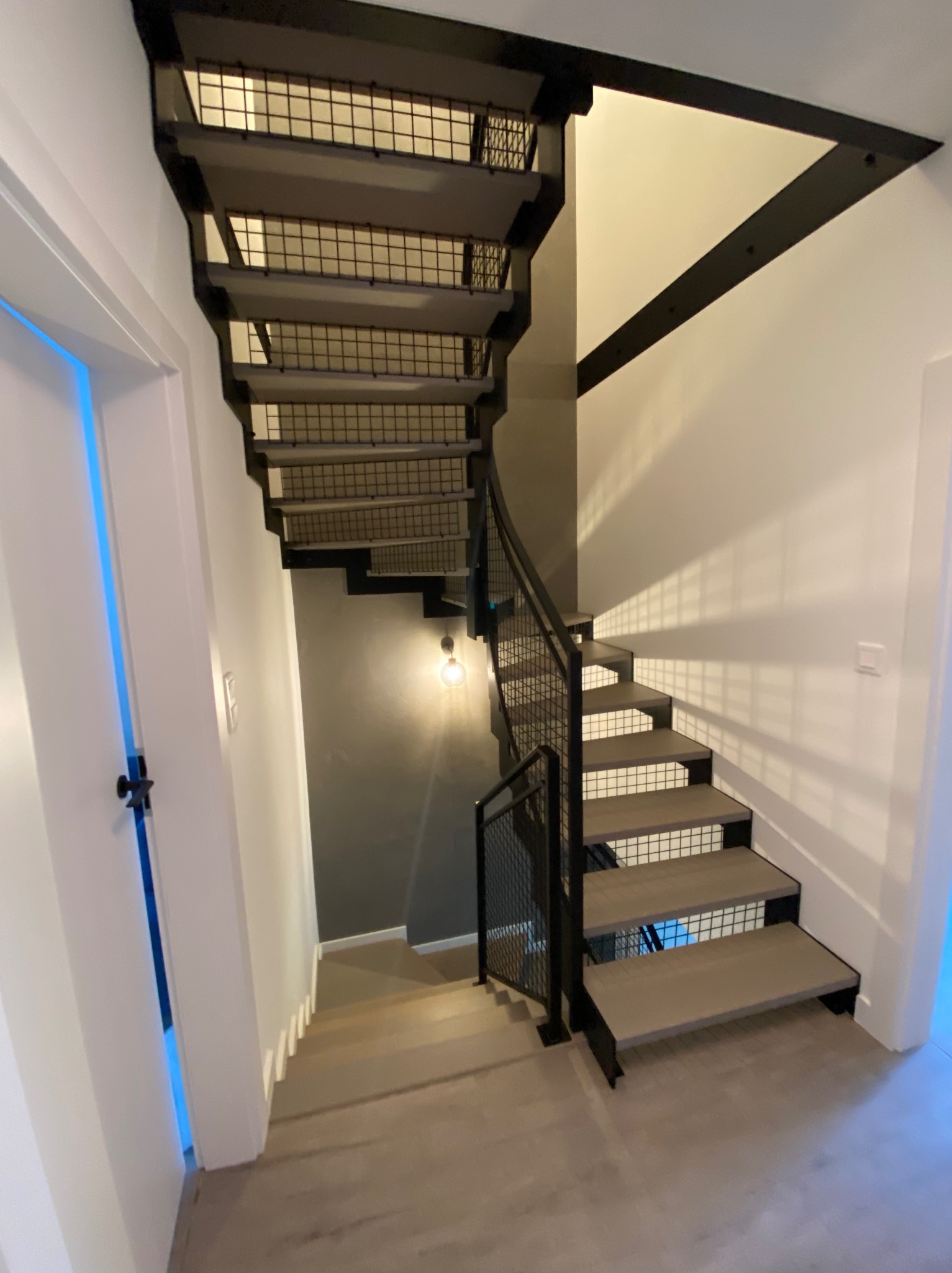 Nowoczesne schody z metalową balustradą i drewnianymi stopniami, oświetlone wiszącą żarówką, widok z dołu.