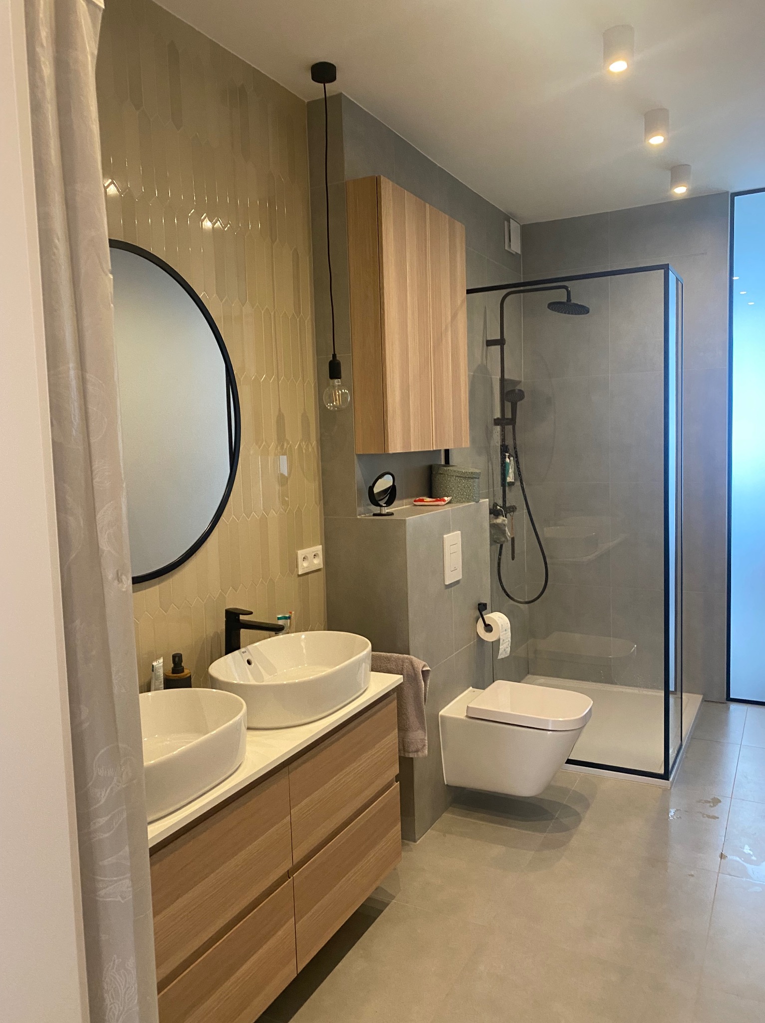 Nowoczesna łazienka z dwoma umywalkami nablatowymi, kabiną prysznicową typu walk-in z czarną armaturą, wiszącą toaletą i szafką z drewnianymi frontami.