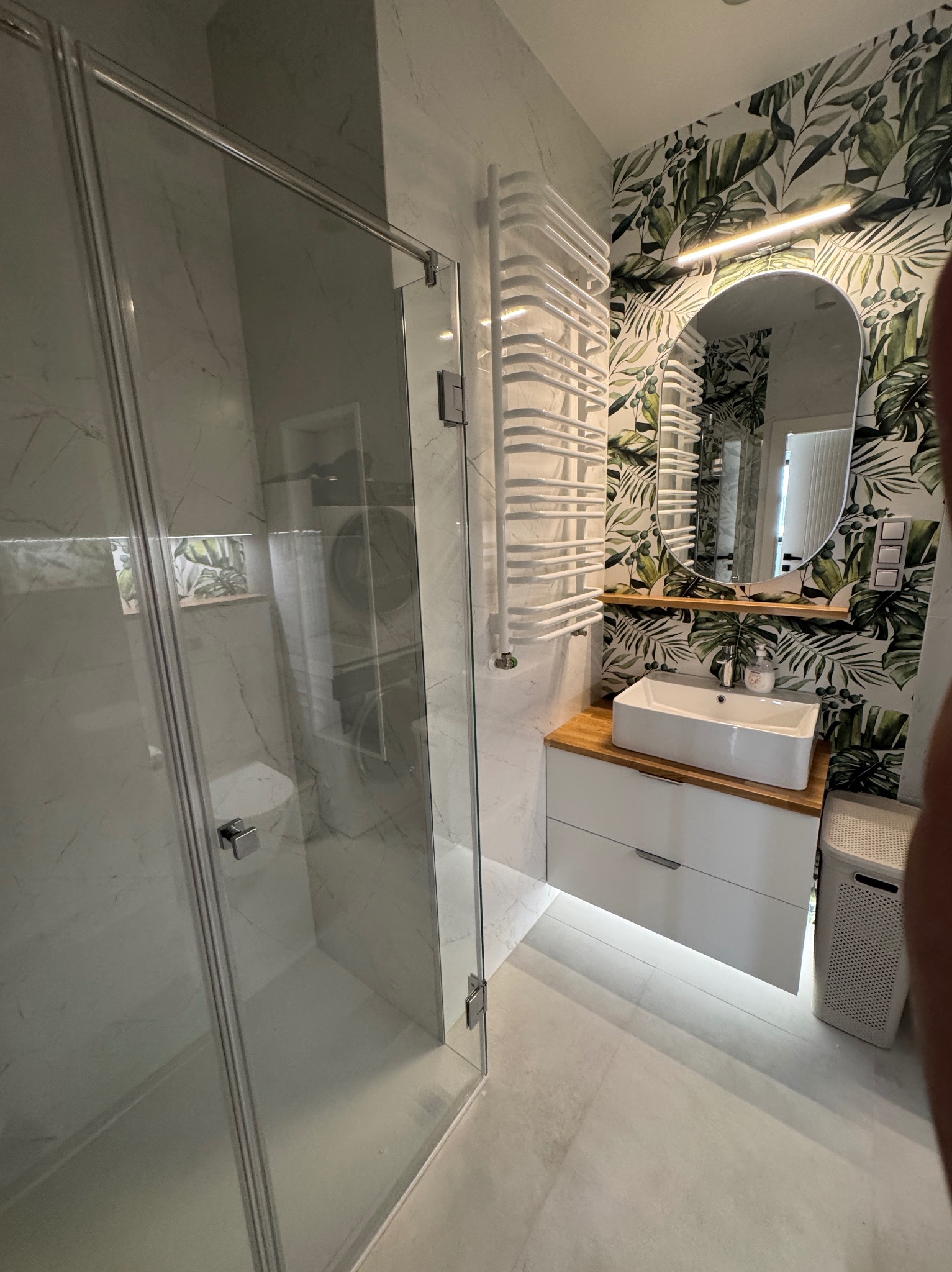 Nowoczesna łazienka z kabiną prysznicową typu walk-in, umywalką na szafce z drewnianym blatem i grzejnikiem drabinkowym na tle ściany w tropikalne liście.