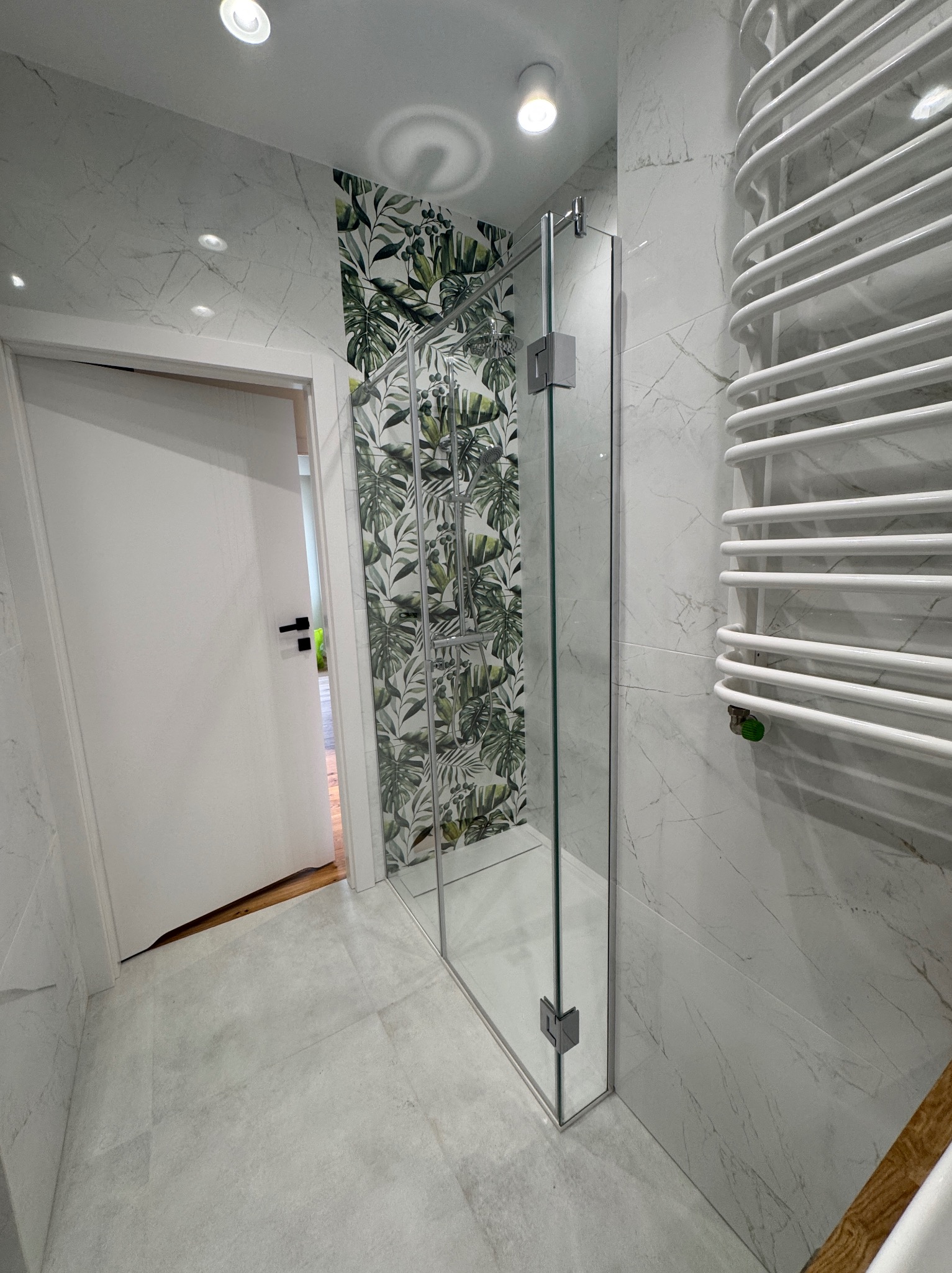 Nowoczesna łazienka po remoncie: kabina prysznicowa ze szklanymi drzwiami, białe marmurowe płytki i grzejnik drabinkowy, akcent tropikalnej tapety.