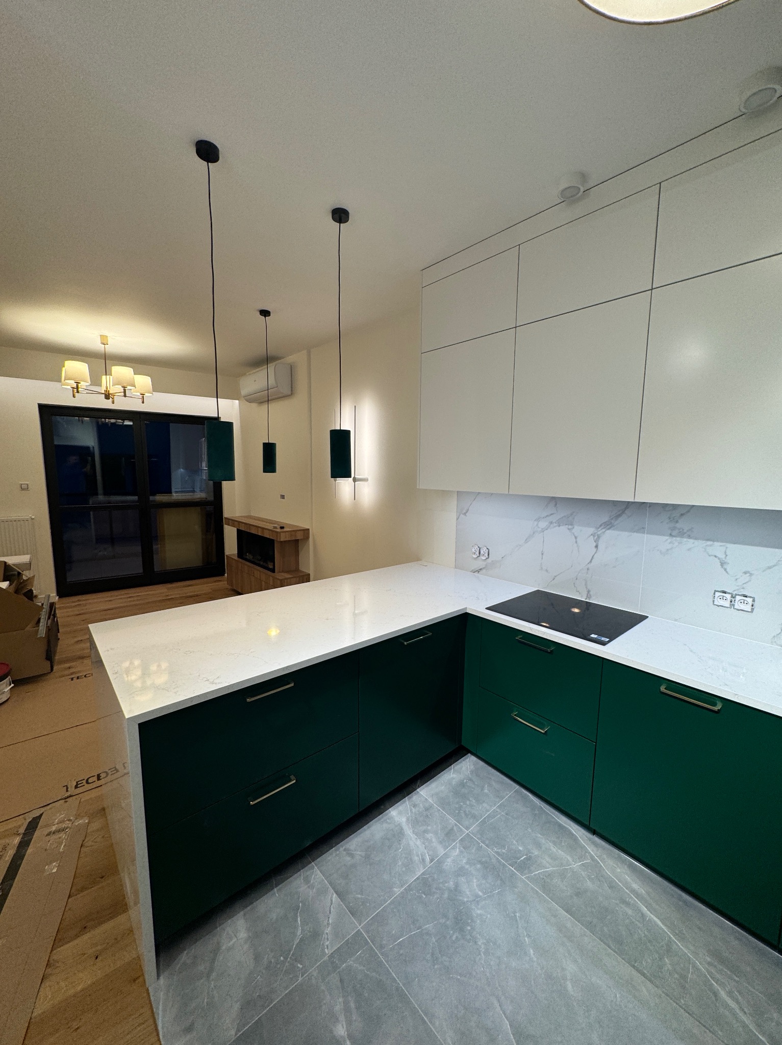 Nowoczesna kuchnia z zielonymi szafkami, białym blatem i marmurową ścianą. Nad blatem wiszą minimalistyczne, czarne lampy. Widoczne szare płytki podłogowe.