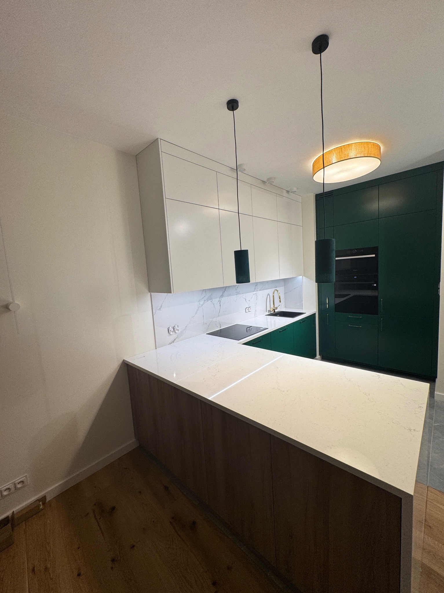 Nowoczesna kuchnia z białymi i zielonymi szafkami, marmurowym blatem i złotą armaturą. Widoczne wiszące lampy i drewniana podłoga. Nietypowe połączenie kolorów.