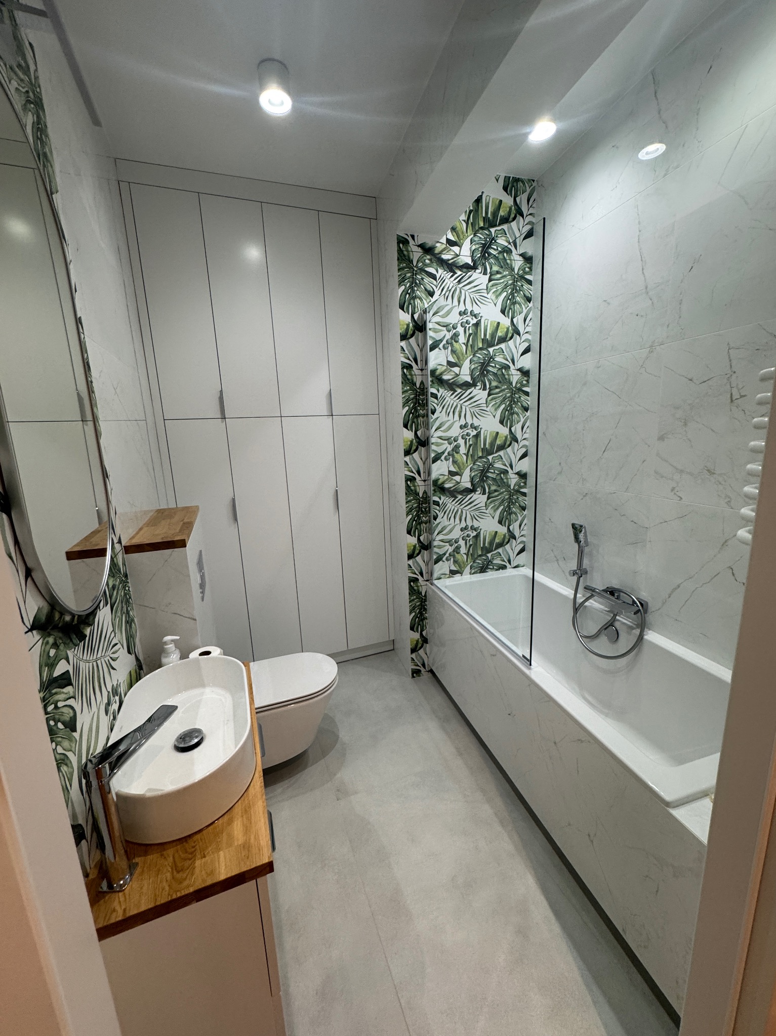Nowoczesna łazienka z wanną w obudowie z płytek imitujących marmur, umywalką nablatową na drewnianym blacie i szafą w zabudowie, akcent tropikalnych liści na ścianie.