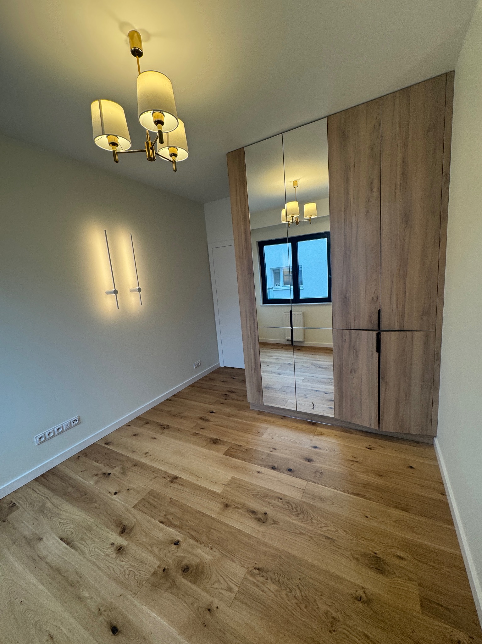 Nowoczesne wnętrze z drewnianą podłogą i szafą w zabudowie z lustrzanymi drzwiami. Widoczny designerski żyrandol i oświetlenie ścienne.