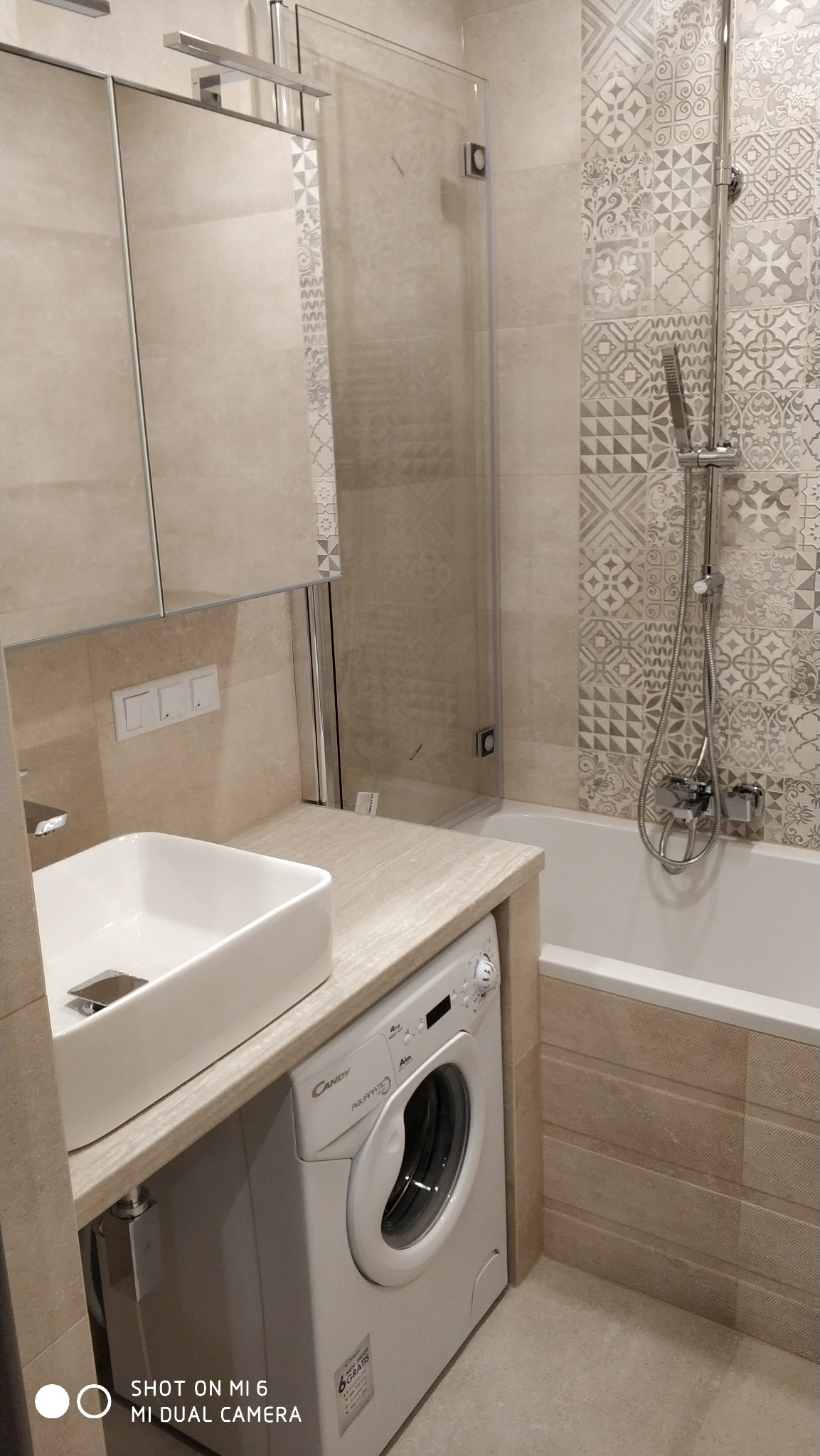 Jasna łazienka po remoncie z umywalką nablatową, pralką pod blatem, kabiną prysznicową i dekoracyjnymi płytkami ceramicznymi w odcieniach szarości i beżu.