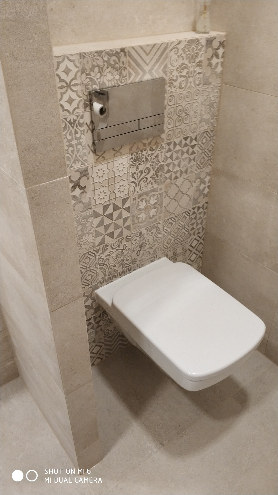 Nowoczesna toaleta podwieszana z mozaiką na ścianie, chromowany przycisk spłukujący z uchwytem na papier toaletowy, beżowe płytki podłogowe i ścienne.