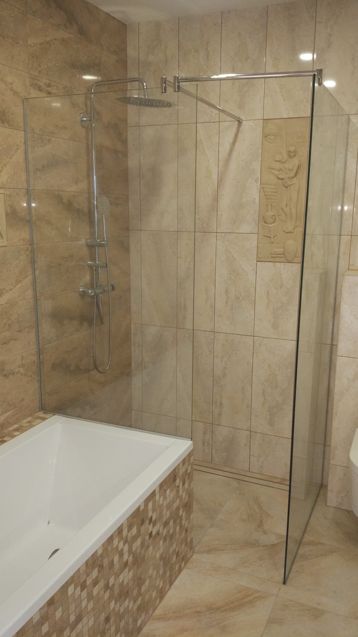 Nowoczesna łazienka z beżowymi płytkami imitującymi marmur, kabina prysznicowa typu walk-in z deszczownicą, wanna obudowana mozaiką i dekoracyjny płaskorzeźba na ścianie.