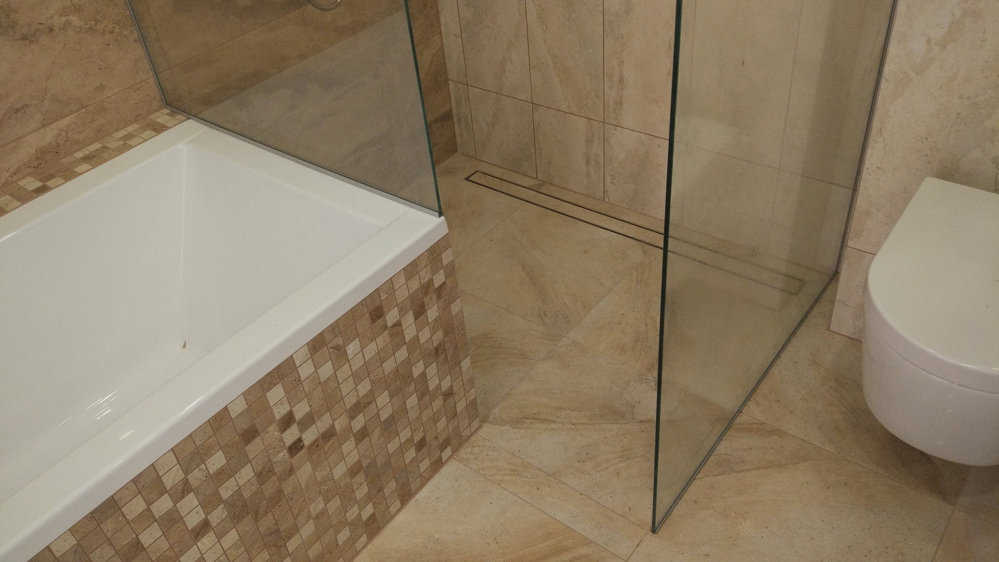 Nowoczesna łazienka z beżowymi płytkami, kabiną prysznicową typu walk-in z odpływem liniowym, wanną obudowaną mozaiką i podwieszaną toaletą.