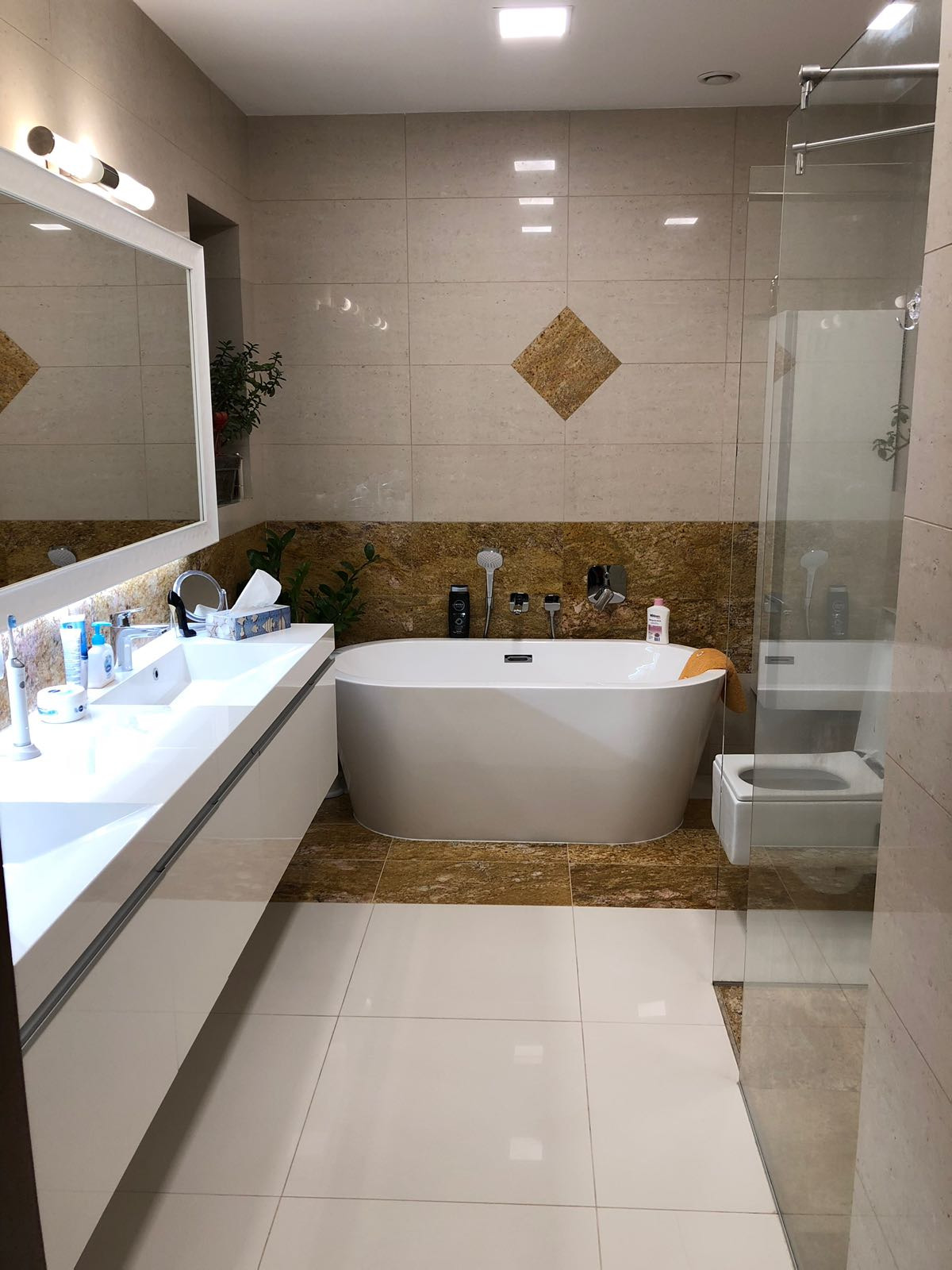 Nowoczesna łazienka z wolnostojącą wanną, umywalką nablatową i kabiną prysznicową typu walk-in, wykończona beżowymi i brązowymi płytkami imitującymi kamień.