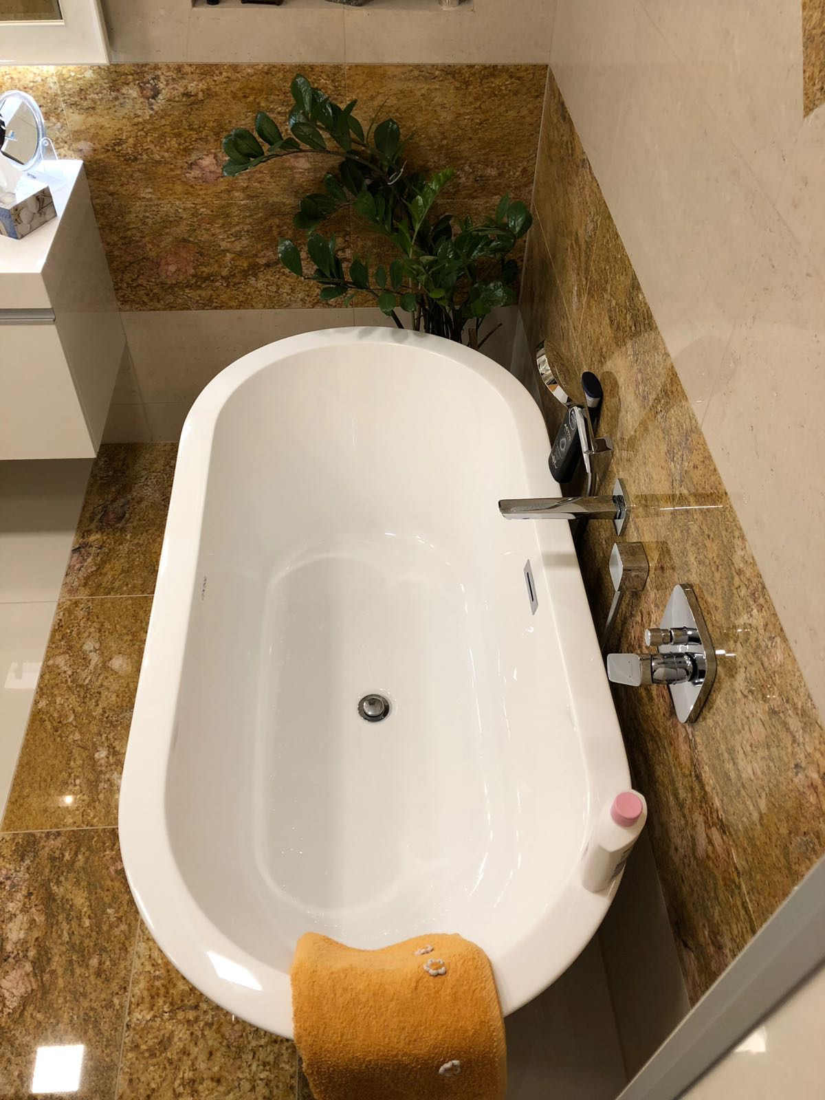 Biała, owalna wanna z chromowaną armaturą na tle ściany wyłożonej płytkami imitującymi marmur, uzupełniona rośliną doniczkową i pomarańczowym ręcznikiem z dekoracyjnymi przeszyciami.